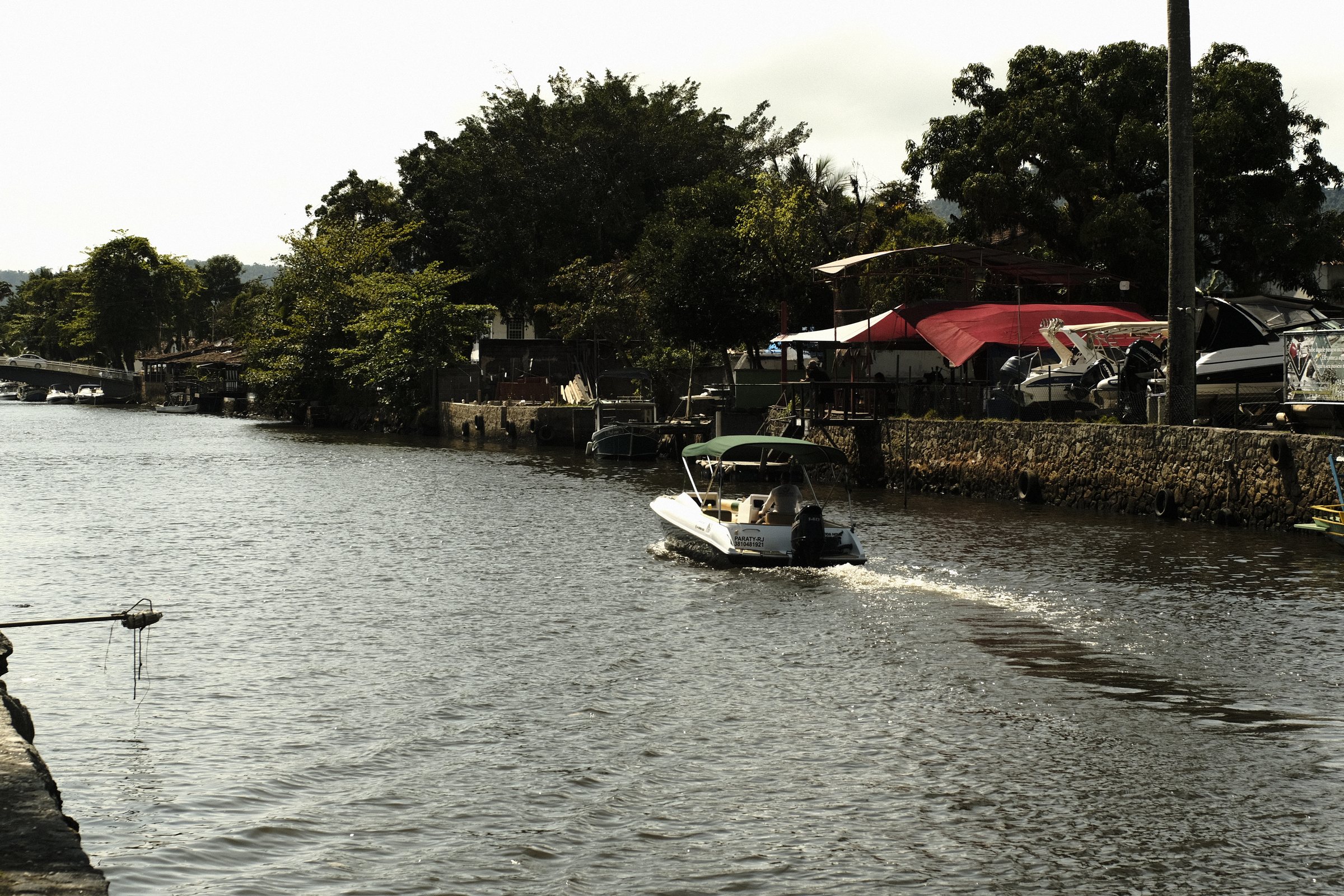 Small motorboat crossing the canal in Paraty, with trees and boat sheds along the bank