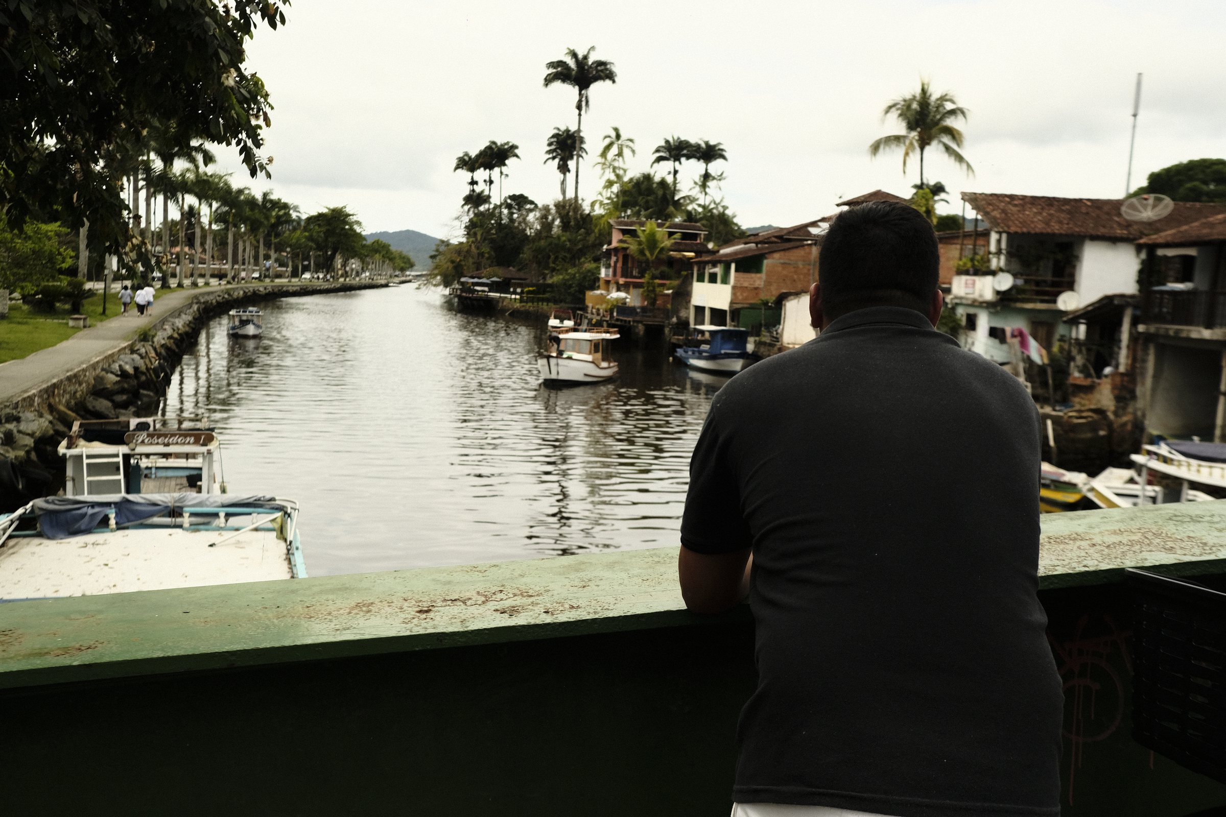 Man leaning on a bridge railing, looking over a canal with moored boats and palm trees in Paraty